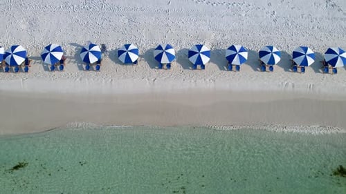 Drone aerial view of blue and white umbrellas on a white sand beach with clear waters on a sunny day