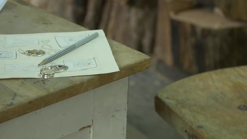 Wooden table in a workshop with drawings and jewelry on it, man walking in the background
