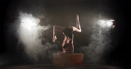 Un contorsionista flexible haciendo pose de escorpión en una caja entre focos y humo