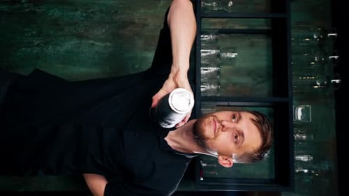 Vertical Video Professional Bartender in Black Shirt Holds in Hands Steel Shaker Blurred Bottles on