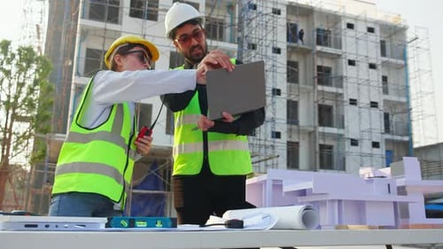 Team architect and builder talking and holding laptop looking model house at construction site.