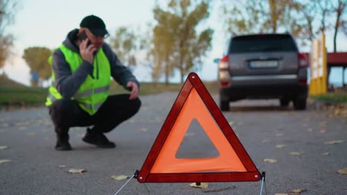 Red Warning Triangle on Background of Blurred Road with Broken Car and Driver in Reflective Vest