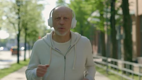 Portrait of the Mature Man Runner is Jogging in the Street Looking at the Camera Wearing Headphones