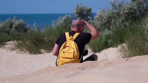 Man Tourist with Backpack Sits on Sand Beach Dune Looking at Amazing Sea View