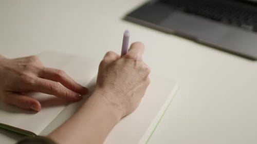 Woman Writes in Notebook at Desk with Laptop