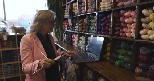 Female Business Owner Taking Inventory At Laptop In Yarn Store