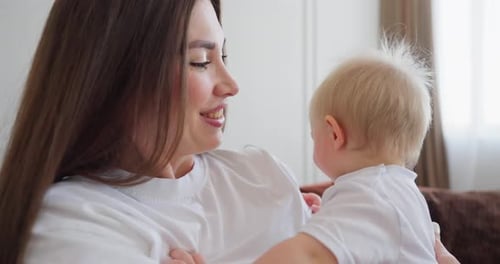 Junge Mutter kuschelt und spielt mit ihrem lächelnden Baby auf einem gemütlichen Sofa in einem hellen Wohnzimmer