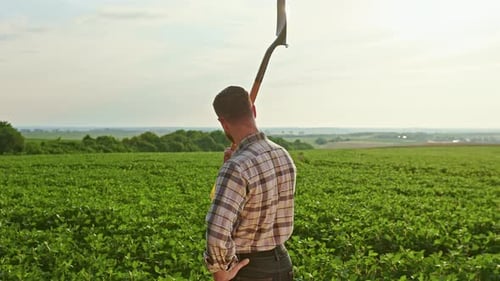 Back View of a Farmer Wearing a Plaid Shirt Standing on the Field A Man Raises a Spade Looking at