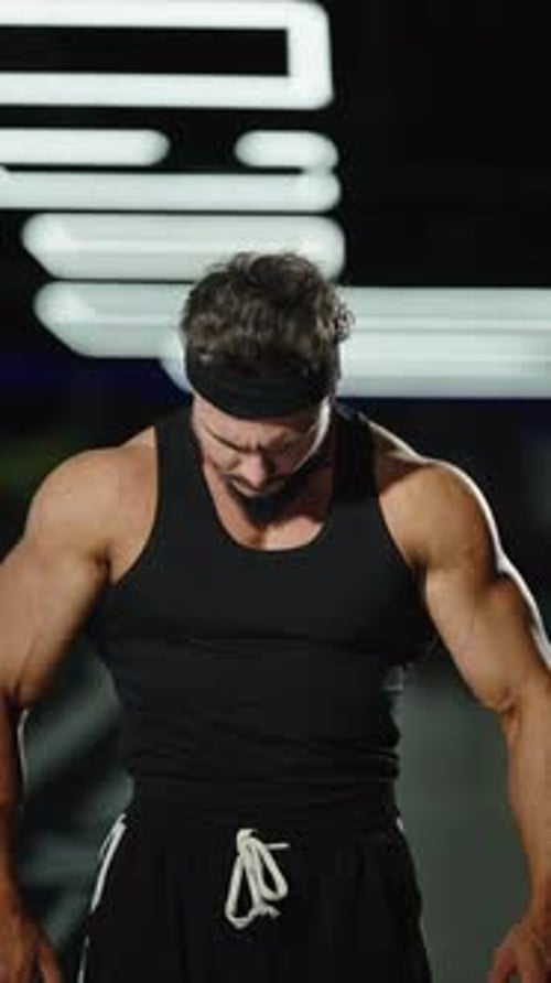 Focused Muscular Athlete in a Black Tank Top and Headband Under Fluorescent Gym Lights Showcasing