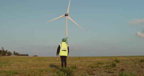 A technician or engineer wearing a helmet and a reflective vest walks towards a wind turbine. In