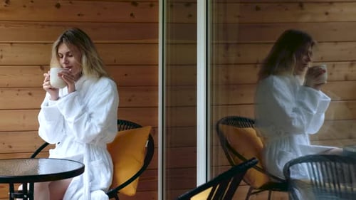 mulher em um roupão de banho bebe chá na varanda perto da janela com seu reflexo