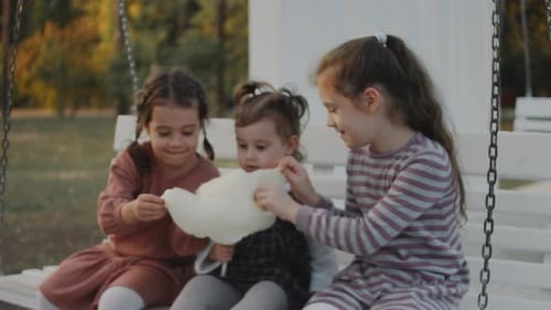 Three Little Girls Having Fun on a White Swing in a Quiet Treelined Park Background Sisters Eating
