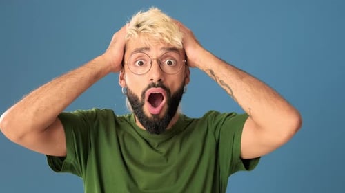 Shocked young man looking at camera covering head with palms, isolated on blue background in studio