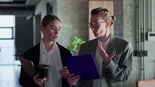Two Businesswoman Girls in Business Clothes and Jackets Walk Along the Corridor in a Modern Office