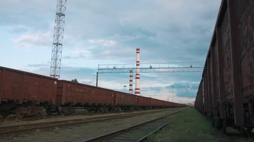 View of Freight Trains at a Railway Yard with Metal Structures and Chimneys Against a Cloudy Sky