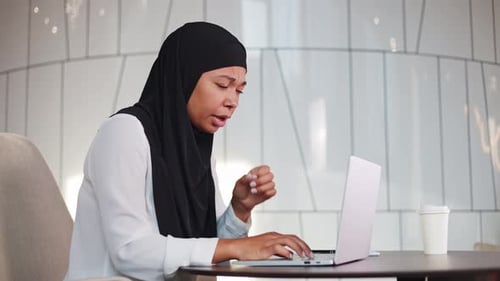 Unhappy Multicultural Woman in Hijab Coughing While Working on Office Laptop