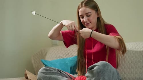 Woman Sits Knitting with Red Yarn on Couch