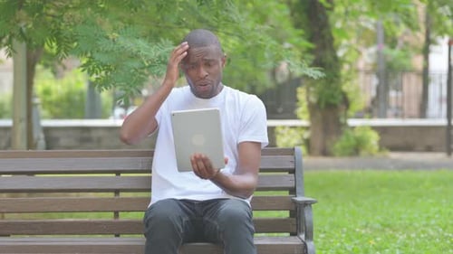 African American Man having Trade Loss on Tablet while Sitting on Bench in Park