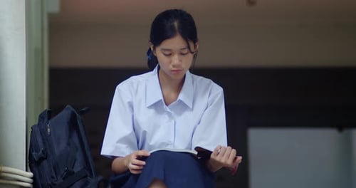 A cute Thai or Asian high school girl in a white school uniform reading a book for exam preparation,