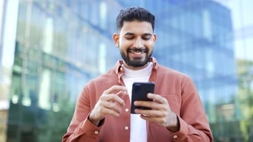 Smiling businessman uses a mobile phone while standing on the street near a modern office building.