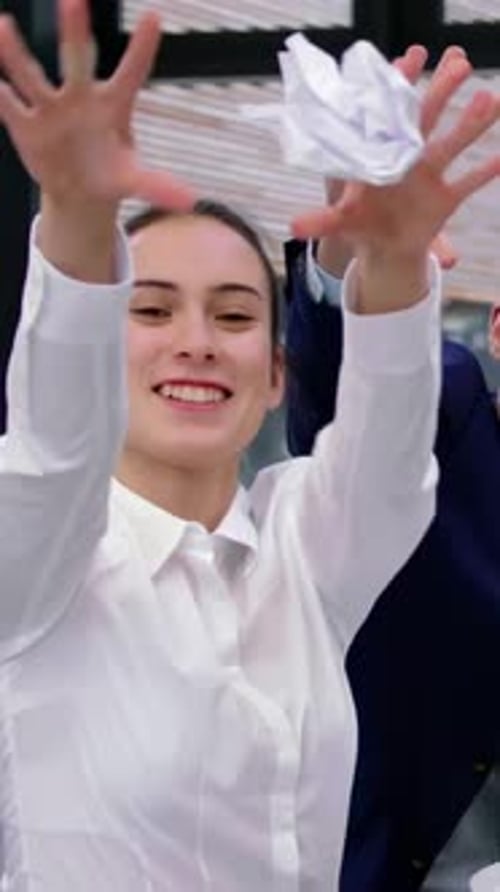 Coworkers tossing crumpled paper balls at office window, woman wearing white blouse, navy suit