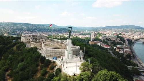 Liberty statue overlooking budapest cityscape with hungarian flag waving in the citadel, drone orbit