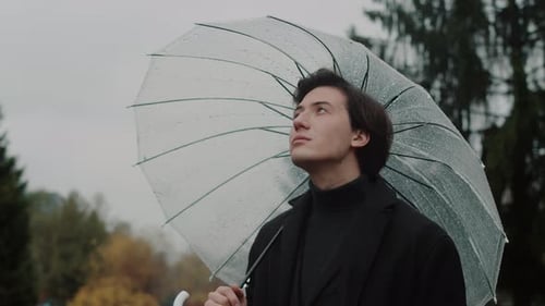 Young Man in a Coat Standing Under a Transparent Umbrella in the Autumn City Park Looking Up at