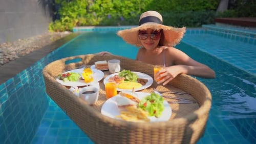 A young woman tourist has her own personal breakfast on a floating table in a private swimming pool.