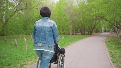 Rear View Caregiver Pushing Wheelchair Down Paved Park Path Lined With Trees Steady Forward Motion
