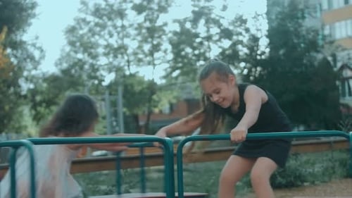 Zwei süße, fröhliche kleine Mädchen reiten auf Merrygoround auf dem Spielplatz und haben Spaß an einem sonnigen Sommer