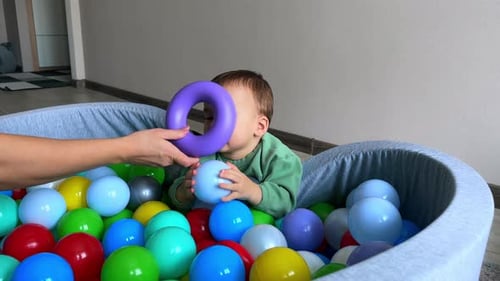 Happy Caucasian boy lay on the wall of the soft dry basin. Mom’s hand gives a toy ring to her son.