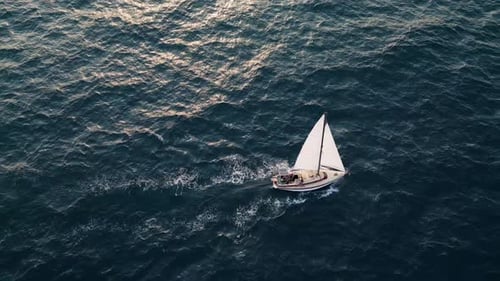 Luftbild eines weißen Segelboots, das alleine auf tiefblauem Meer segelt. Ruhige Wellen schimmern unter Sonnenlicht.