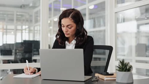 Businesswoman Working on Laptop and Taking Notes in Notebook at Office Desk