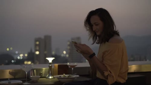 Young Woman with Smartphone Drinking Cocktail Sitting on Terrace in Bar At