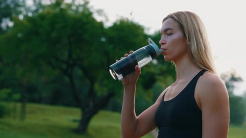 Side View of the Caucasian Runner Woman Standing in the Park Drinking Water From the Sport Bottle