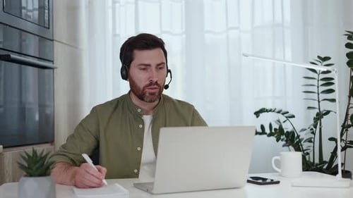 Freelancer in Headset Taking Notes During Video Call on Laptop at Home Office