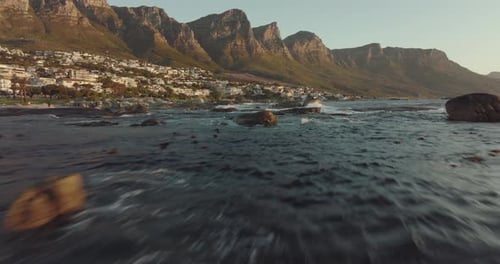 Drone flies just over the sea at camps bay in cape town south africa - wavy sea, many houses on a hi