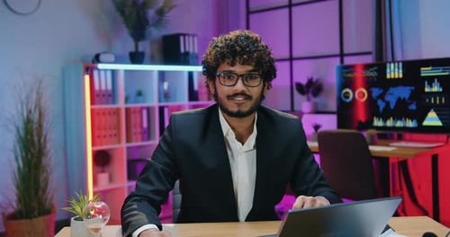 Attractive business man in glasses sitting in front of camera in evening office with satisfied face