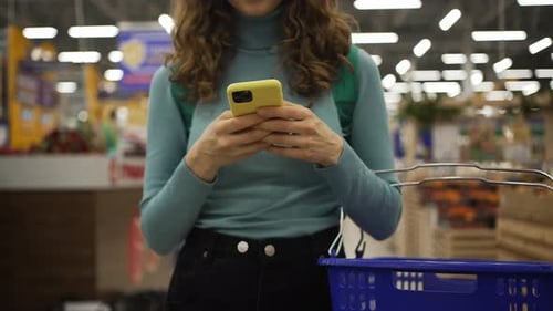 Woman Using Smartphone and Holding Shopping Basket in Supermarket
