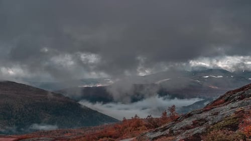 Heavy stormy clouds backlit by t the sun whirl above the autumn valley casting dark shadows. The fog