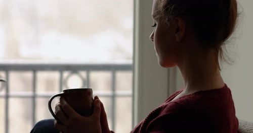 Young woman quietly watches snowflakes fall outside while enjoying hot beverage in mug. 4K