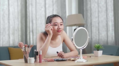Asian Woman Looks In The Mirror Combing Long Black Eyelashes With Mascara Brush And Smiles