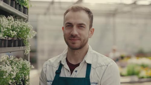 Man Smiling Beside Potted Flowers in Greenhouse