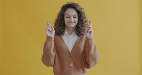 Portrait of Young Student Making Wish Crossing Fingers and Closing Eyes Smiling on Yellow Background