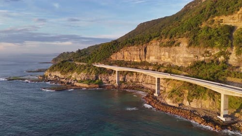 Luftaufnahmen der Sea Cliff Bridge, mit einem Fahrzeug, das auf der kurvenreichen Straße über dem blauen Weg navigiert