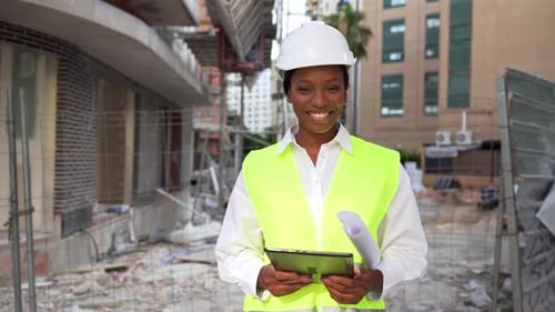 African American architect in helmet and vest holding tablet and blueprint on site smiling at camera