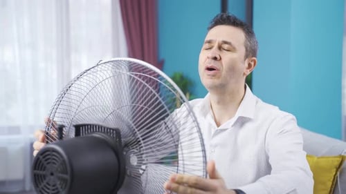 Overwhelmed by the heat, the man turns on the fan and relaxes.