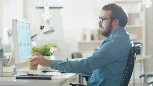 Junger bärtiger Mann macht sich Notizen, während er am Desktop-Computer arbeitet. Er nippt an Kaffee. Er sitzt in einer Crea