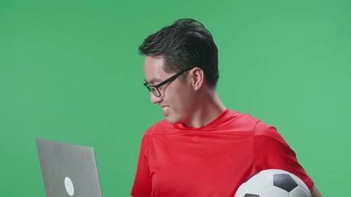 Close Up Side View Of Asian Man With A Ball Watching Soccer Match On A Laptop In The Green Screen