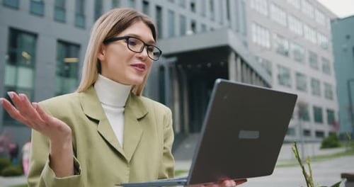 Woman in glasses standing by modern building offices during using a laptop. Virtual meeting